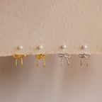 Two pairs of Pearl & Bow Ear Jackets in 18K Gold—one in gold and one in silver—are displayed on a textured beige surface with pearl stud earrings above.