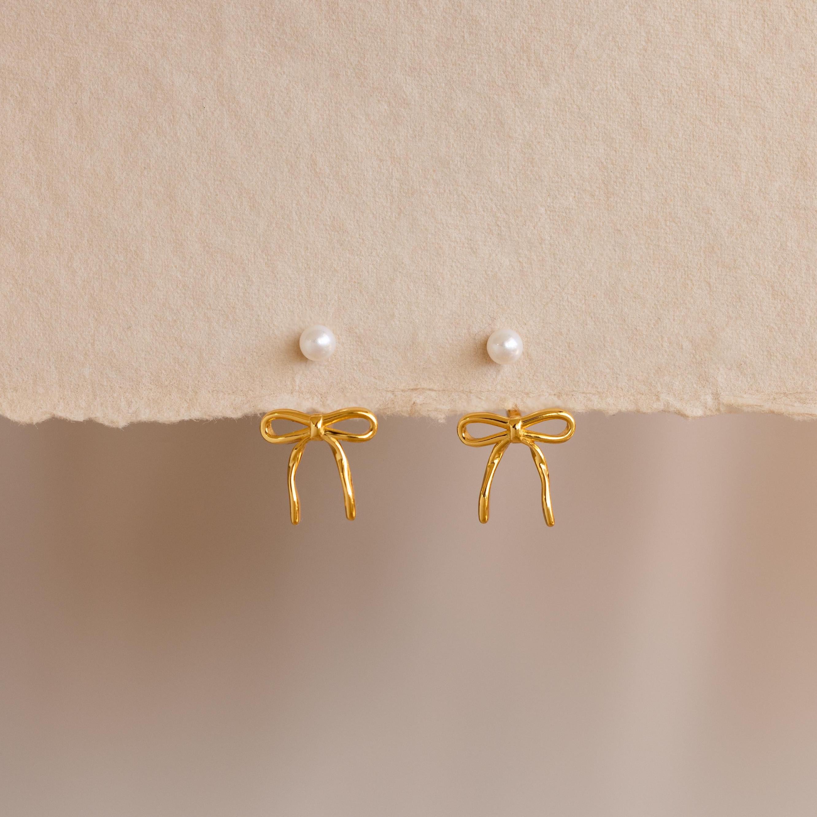 Two Pearl & Bow Ear Jackets, featuring gold bows and small pearl studs, are displayed on a textured beige background—perfect for adding elegance to your look.