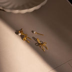 The Pearl & Bow Ear Jackets in 18K Gold, adorned with elegant pearls, rest on a beige surface illuminated by soft, warm light.