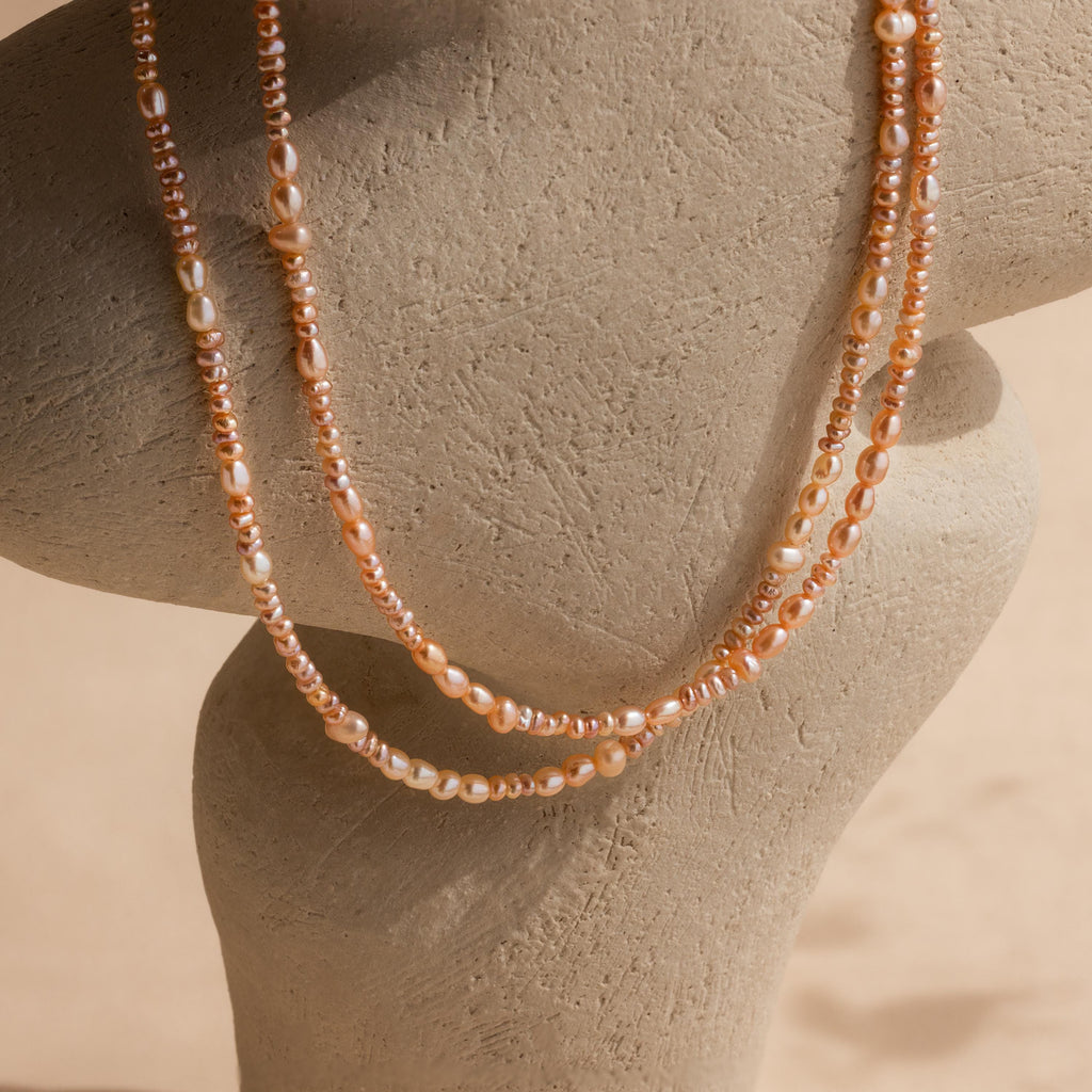 The Blush Pearl Beaded Necklace features two strands of pink freshwater pearls and peach beads, elegantly draped on a beige, textured necklace display bust.