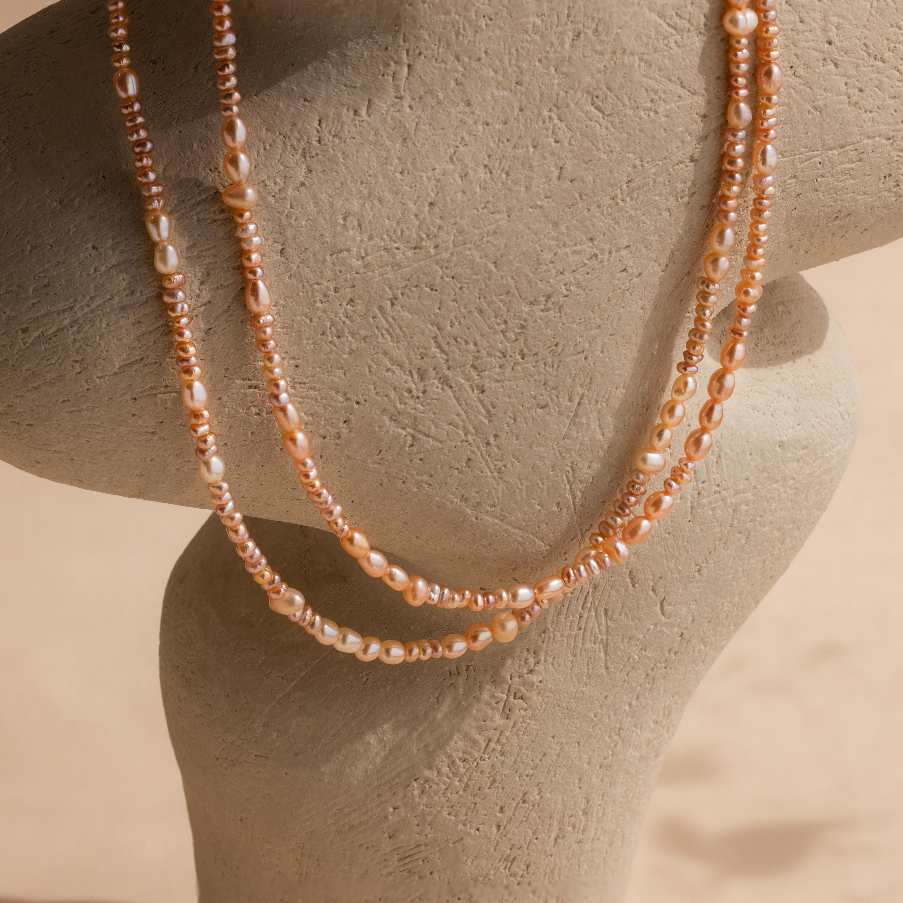 The Blush Pearl Beaded Necklace features two strands of pink freshwater pearls and peach beads, elegantly draped on a beige, textured necklace display bust.