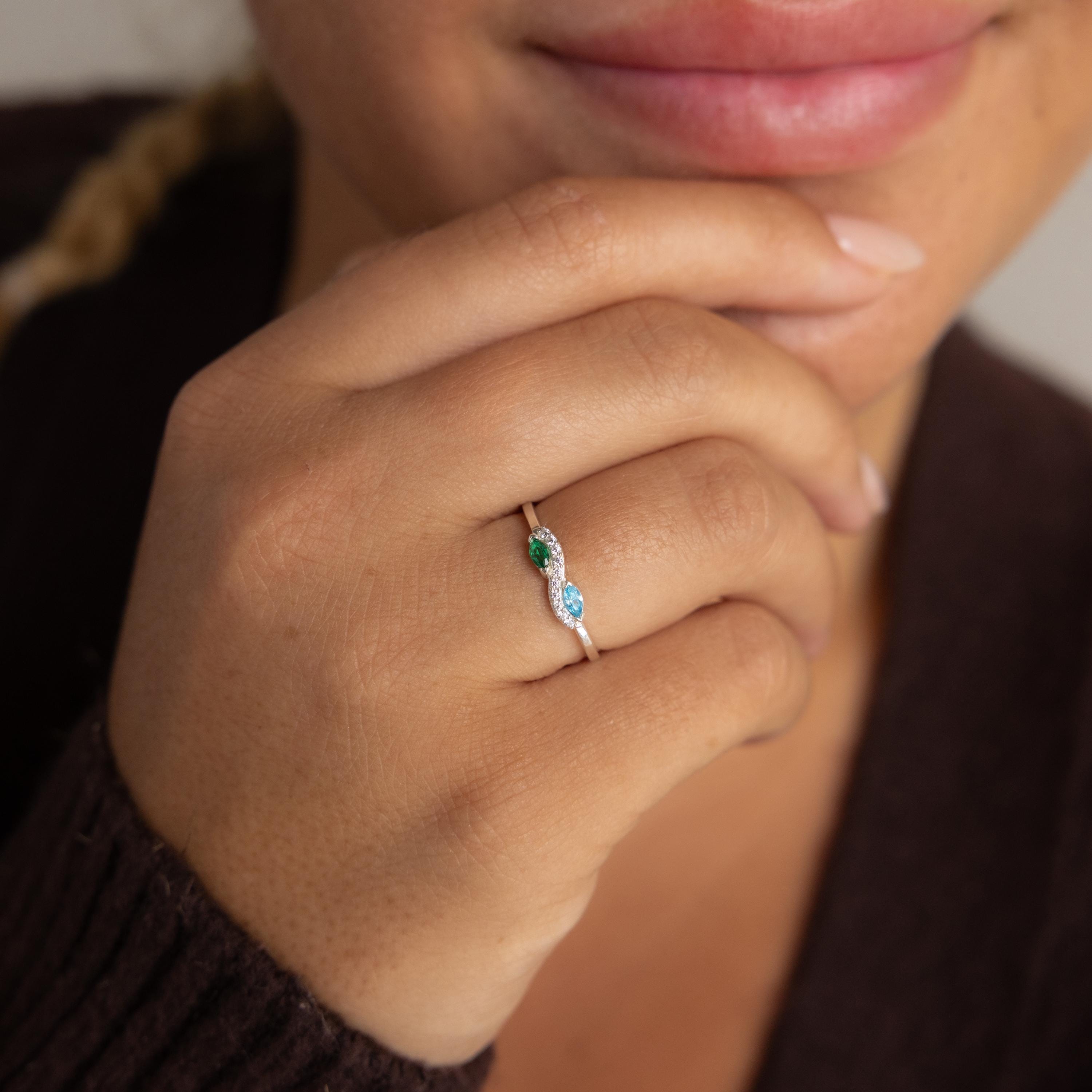 A person wears the Infinity Marquise Birthstone Ring, featuring green and blue stones in an infinity design, with their hand near their lips.