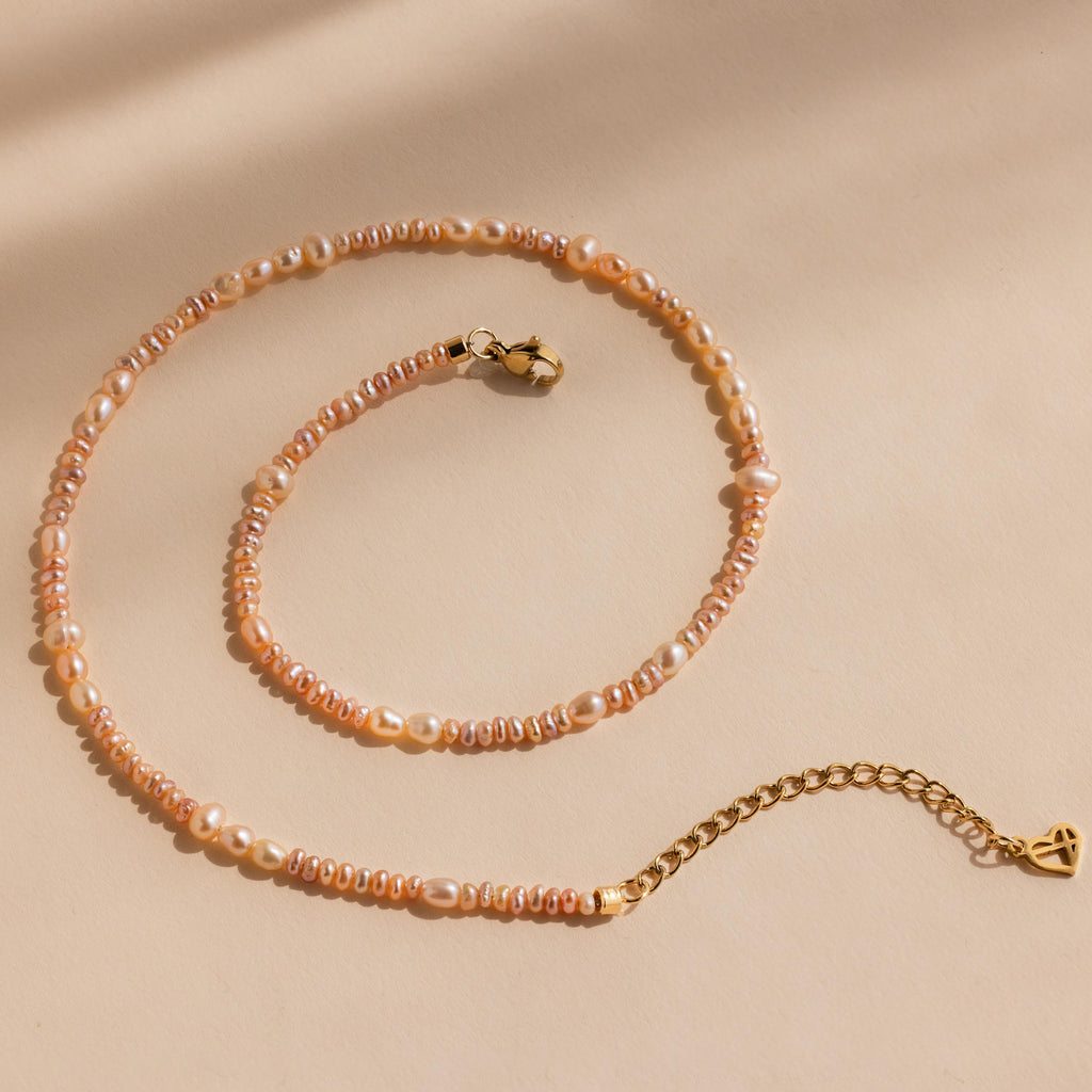 The Blush Pearl Beaded Necklace features pink freshwater pearls with a gold clasp and extender, elegantly displayed on a beige background.
