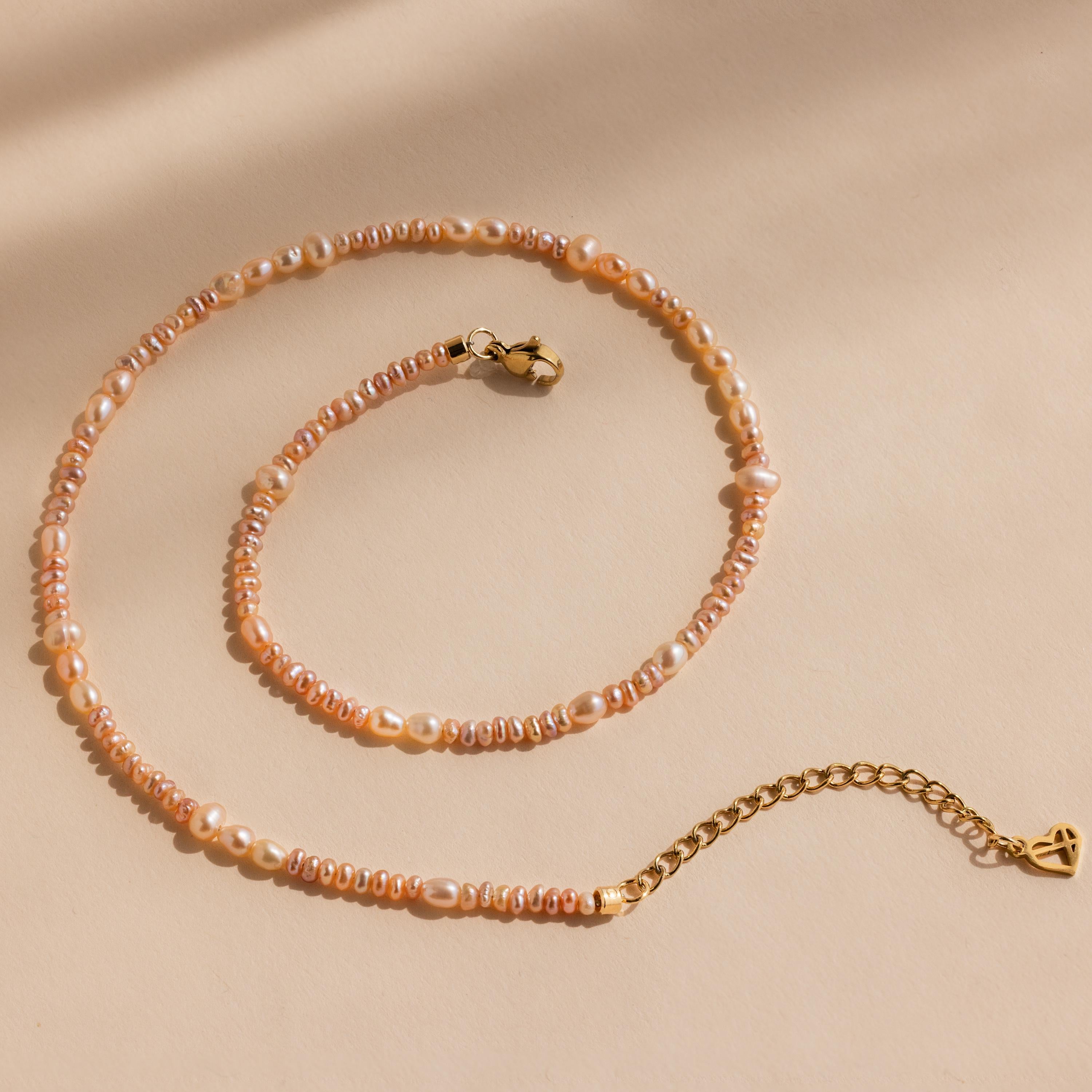 The Blush Pearl Beaded Necklace features pink freshwater pearls with a gold clasp and extender, elegantly displayed on a beige background.