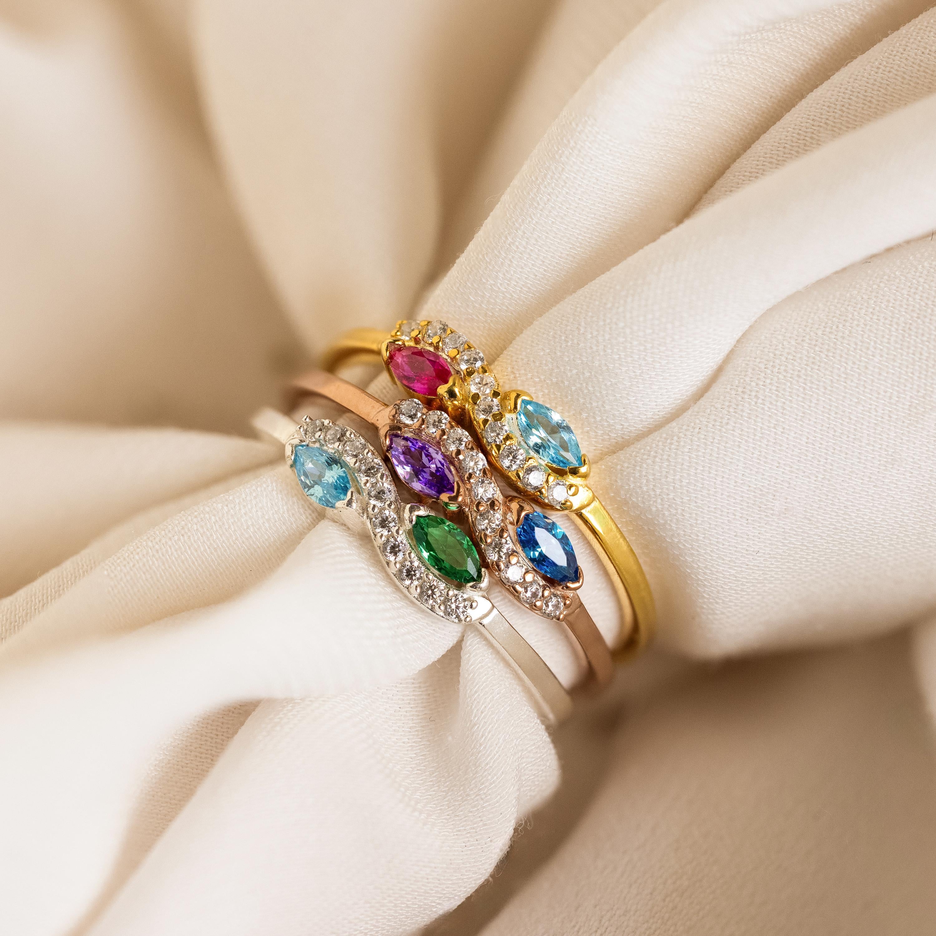 The Infinity Marquise Birthstone Ring, a gold ring with colorful gemstones and small diamonds, is elegantly displayed on cream-colored fabric.