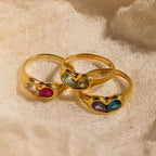 Two Duo Birthstone Heart Signet Rings, each set with colorful gemstones in heart shapes, are displayed on a textured beige fabric—these rings add vibrant charm and a unique pop of color.