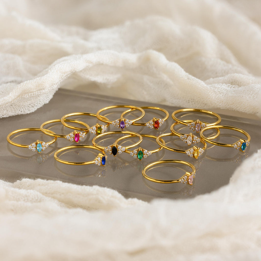 Twelve Tiny Marquise Birthstone Rings with colorful gemstones are arranged on a soft white fabric background.