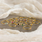 Twelve Tiny Marquise Birthstone Rings with colorful gemstones are arranged on a soft white fabric background.