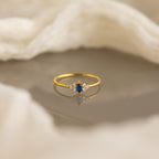 The Tiny Marquise Birthstone Ring features a gold marquise-cut band with a blue gemstone and accent stones, showcased on a reflective surface against soft fabric—a stunning customizable ring for any occasion.