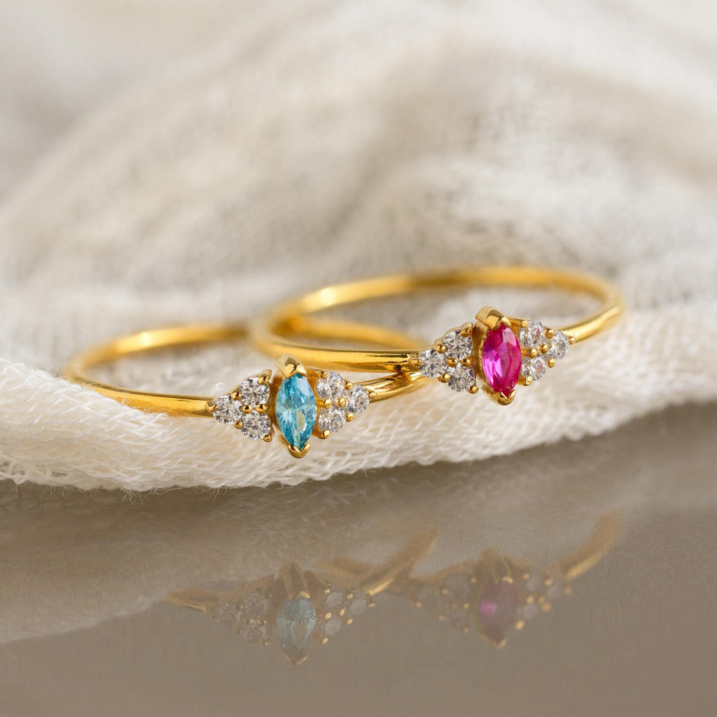 Two Tiny Marquise Birthstone Rings, each featuring marquise-cut blue and pink stones with small clear gems, are elegantly displayed on a white cloth.