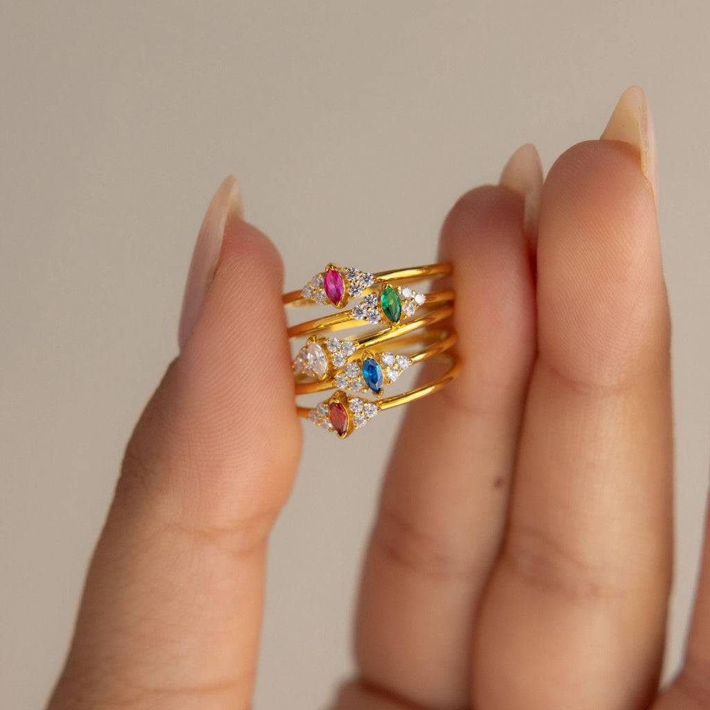 A hand holds three gold rings, including the Tiny Marquise Birthstone Ring featuring colorful gemstones and small diamonds, set against a neutral background.