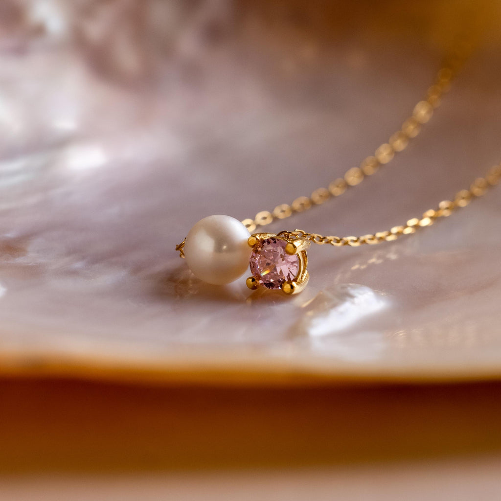 The Pearl & Birthstone Necklace showcases a pearl and pink gemstone pendant, beautifully displayed on a lustrous iridescent shell surface.