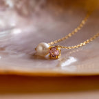 The Pearl & Birthstone Necklace showcases a pearl and pink gemstone pendant, beautifully displayed on a lustrous iridescent shell surface.