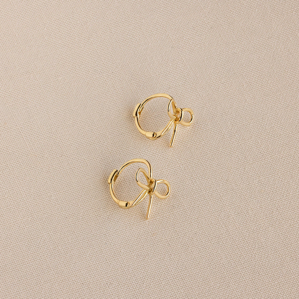 The Classic Ribbon Bow Huggies are small gold hoops with delicate ribbon bow charms, shown on a beige textured fabric background, adding whimsical femininity to any outfit.