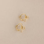 The Classic Ribbon Bow Huggies are small gold hoops with delicate ribbon bow charms, shown on a beige textured fabric background, adding whimsical femininity to any outfit.