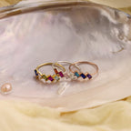 The Multiple Baguette Birthstone Ring, one of three vibrant stackable rings, sits on a shiny iridescent shell with a pearl and soft fabric nearby.