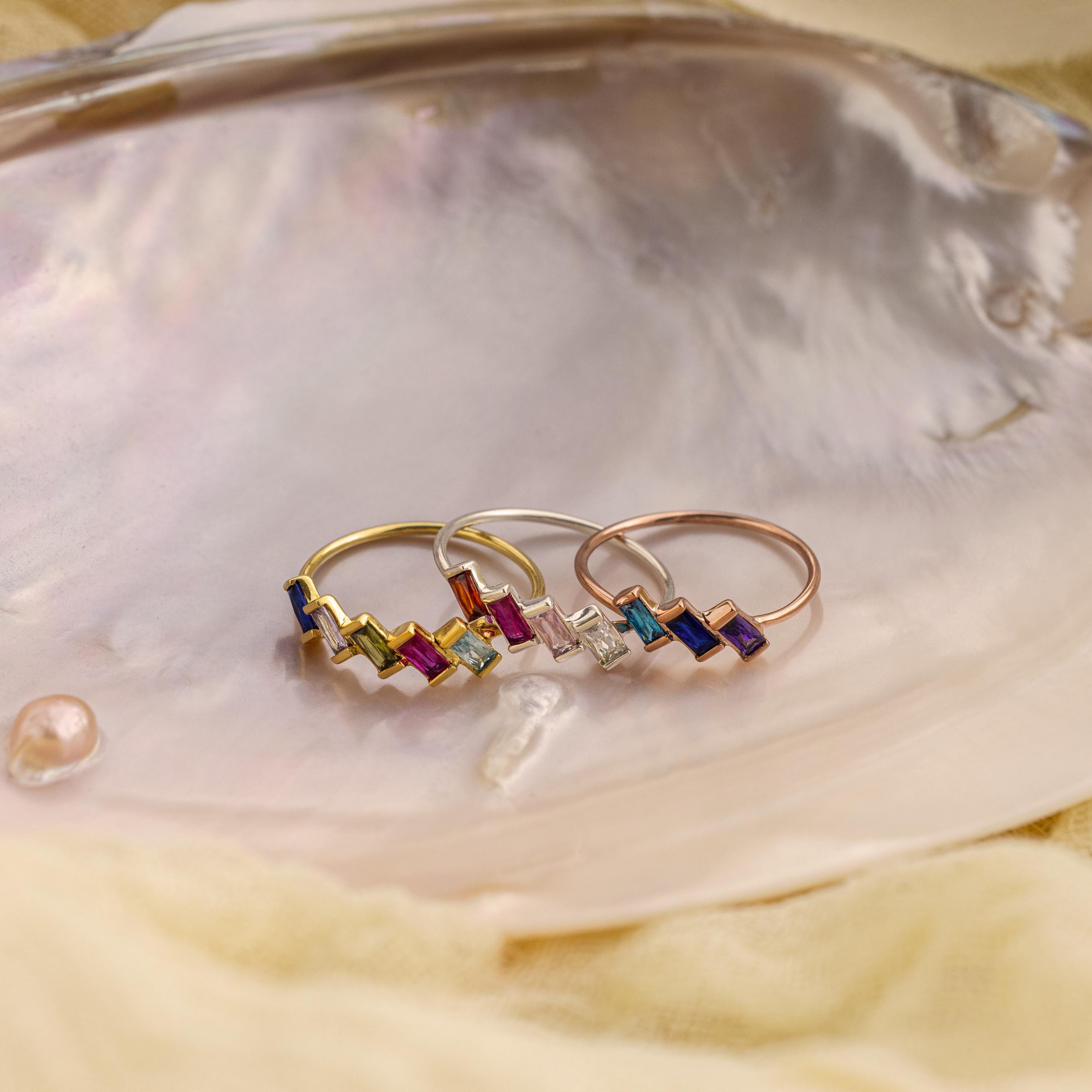 The Multiple Baguette Birthstone Ring, one of three vibrant stackable rings, sits on a shiny iridescent shell with a pearl and soft fabric nearby.