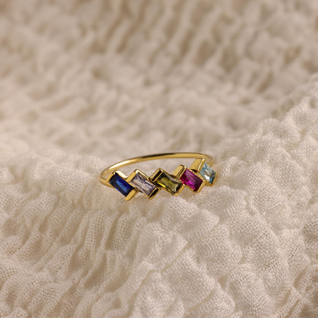 The Multiple Baguette Birthstone Ring features five vibrant rectangular gemstones and sits on textured cream fabric—an elegant, stackable piece for fans of personalized rings.