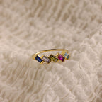 The Multiple Baguette Birthstone Ring features five vibrant rectangular gemstones and sits on textured cream fabric—an elegant, stackable piece for fans of personalized rings.