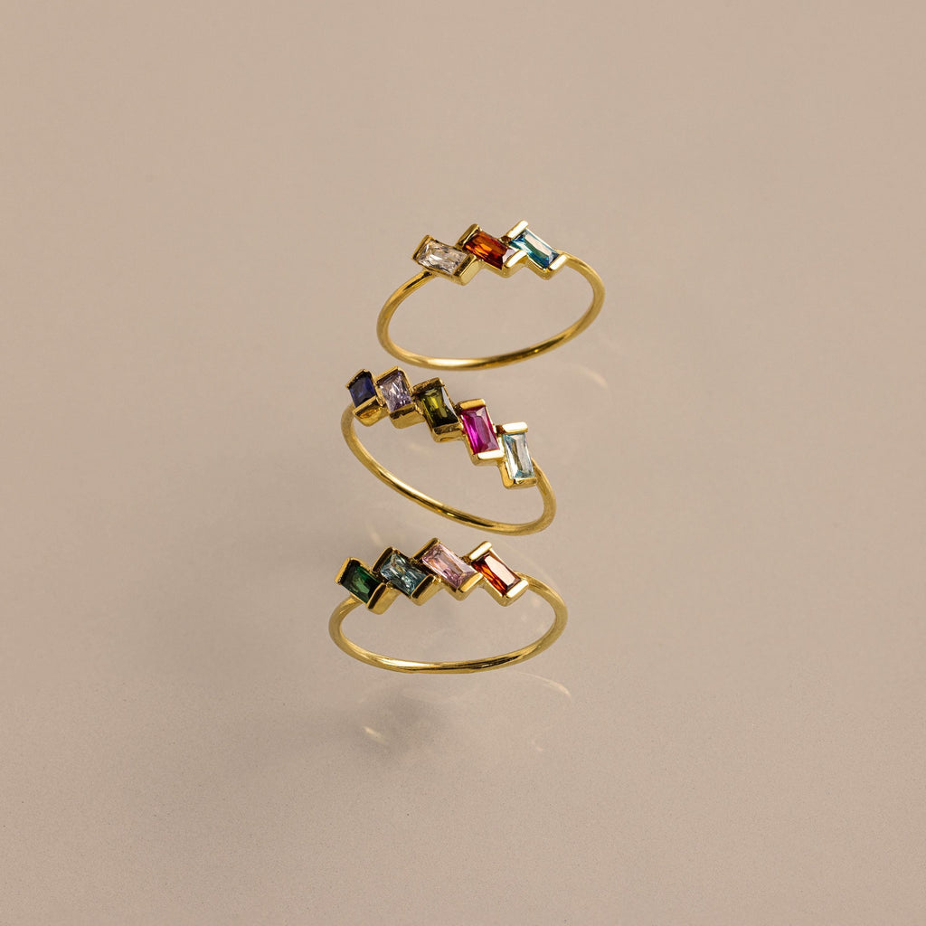 The Multiple Baguette Birthstone Ring is a gold spiral stackable ring with colorful rectangular gemstones, shown on a beige background.
