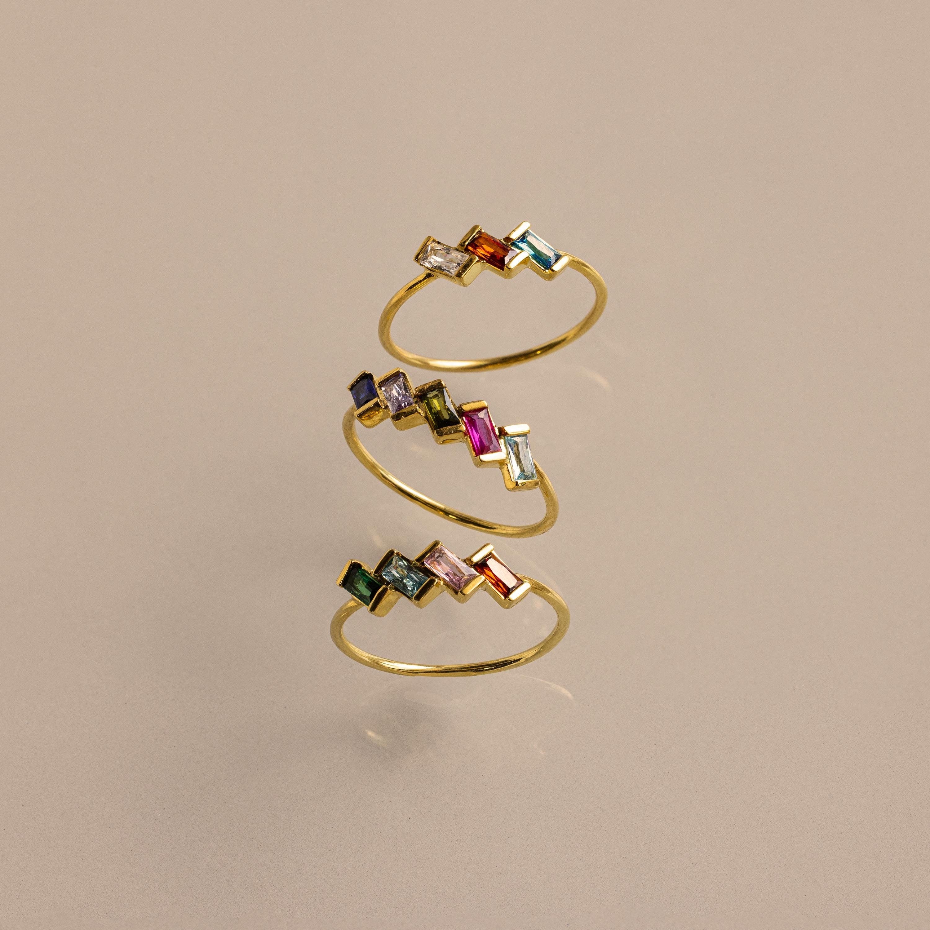 The Multiple Baguette Birthstone Ring is a gold spiral stackable ring with colorful rectangular gemstones, shown on a beige background.