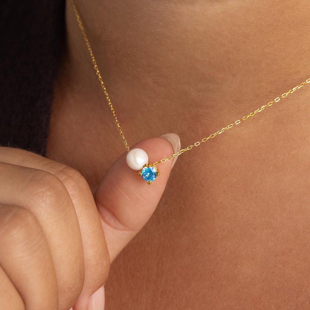 A hand holds the Pearl & Birthstone Necklace, featuring delicate gold pearls and a small blue gemstone pendant—an elegant piece of personalized jewelry perfect for any look.