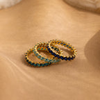 The Eternity Marquise Birthstone Ring features three gold bands with blue, teal, and navy gems on a sandy backdrop—ideal as a customizable birthstone ring or a meaningful Christmas gift.