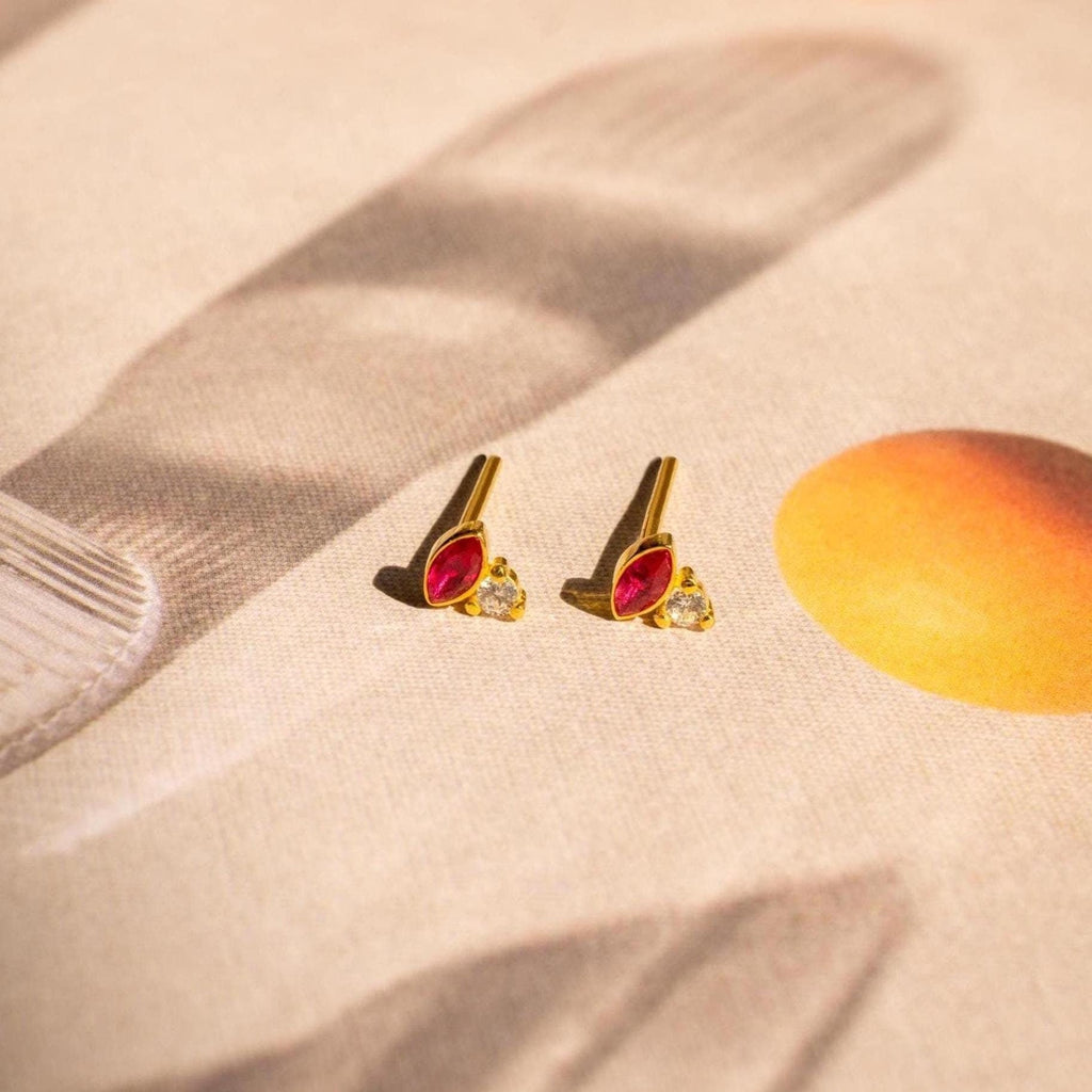 Diamond Marquise Birthstone Studs featuring gold marquise-cut settings with red and clear stones are displayed on a beige background, highlighted by abstract shadows and an orange circle.