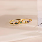 The Cluster Marquise Birthstone Ring, a delicate gold ring adorned with small blue, green, and clear gemstones, is displayed on a smooth beige surface.