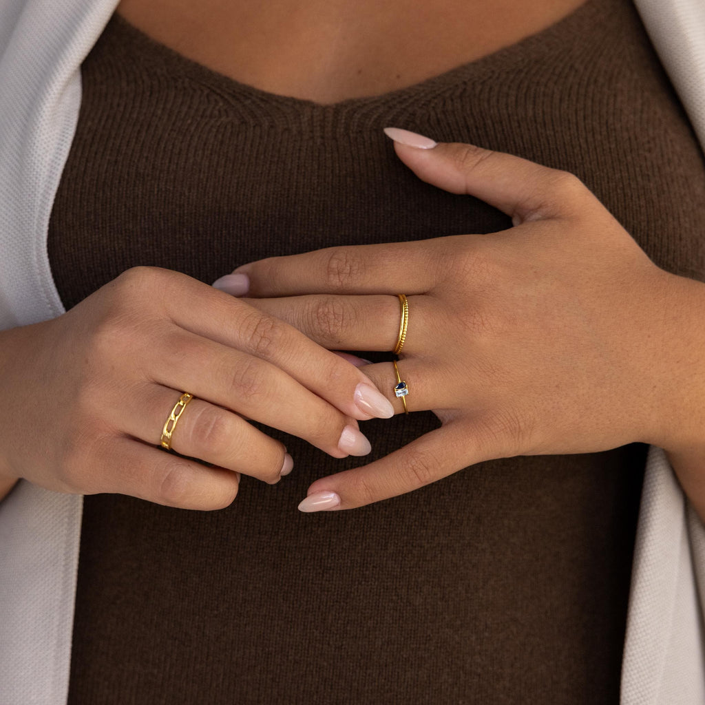 A woman’s manicured hands, adorned with gold rings and the Tiny Toi et Moi Birthstone Ring, rest on a brown top and white cardigan.