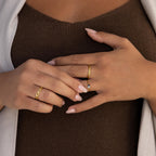 A woman’s manicured hands, adorned with gold rings and the Tiny Toi et Moi Birthstone Ring, rest on a brown top and white cardigan.