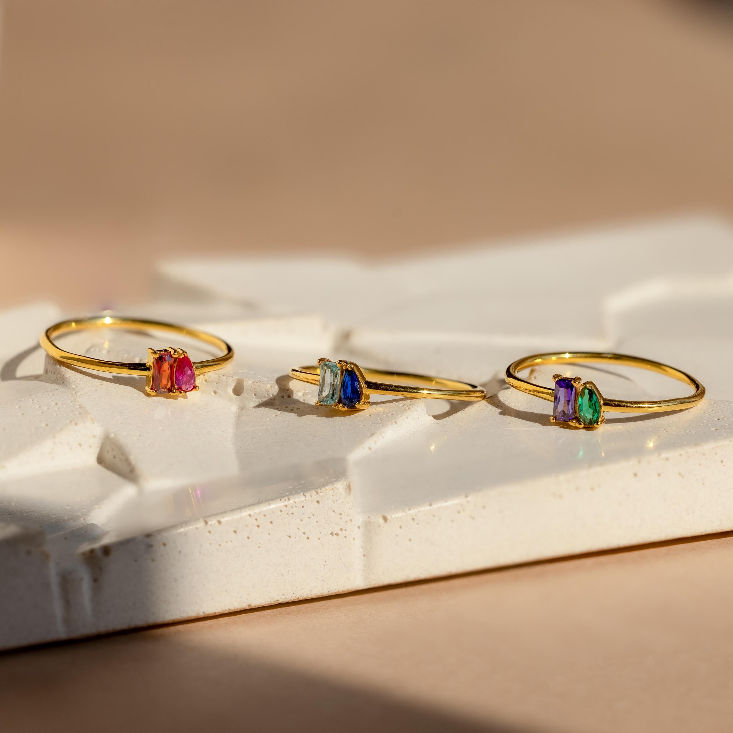 The Tiny Toi et Moi Birthstone Ring, along with two other gold rings featuring colorful gemstones, is displayed on a textured white surface in warm light.