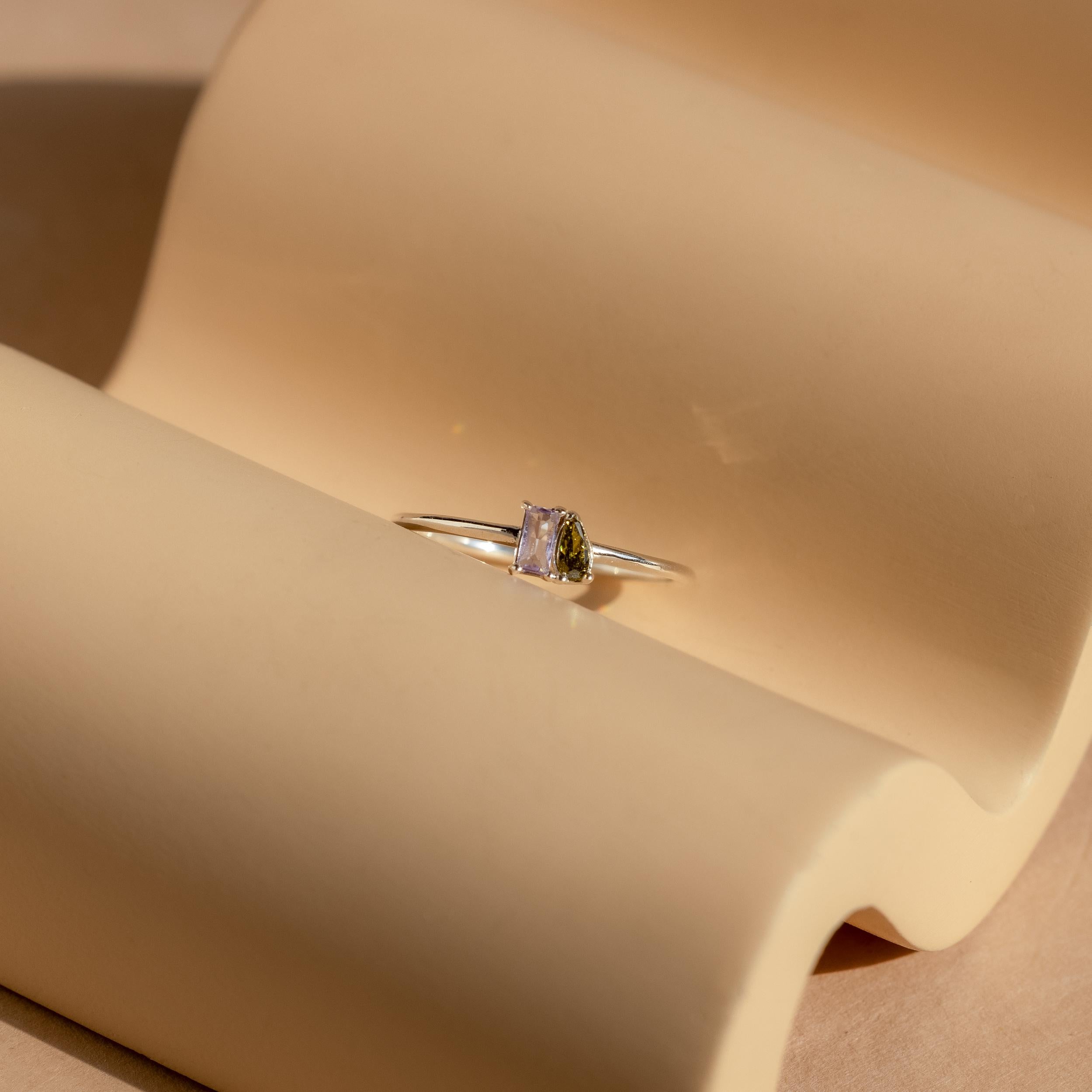 The Tiny Toi et Moi Birthstone Ring showcases a customizable rectangular gemstone, elegantly displayed on a curved beige surface with soft shadows.