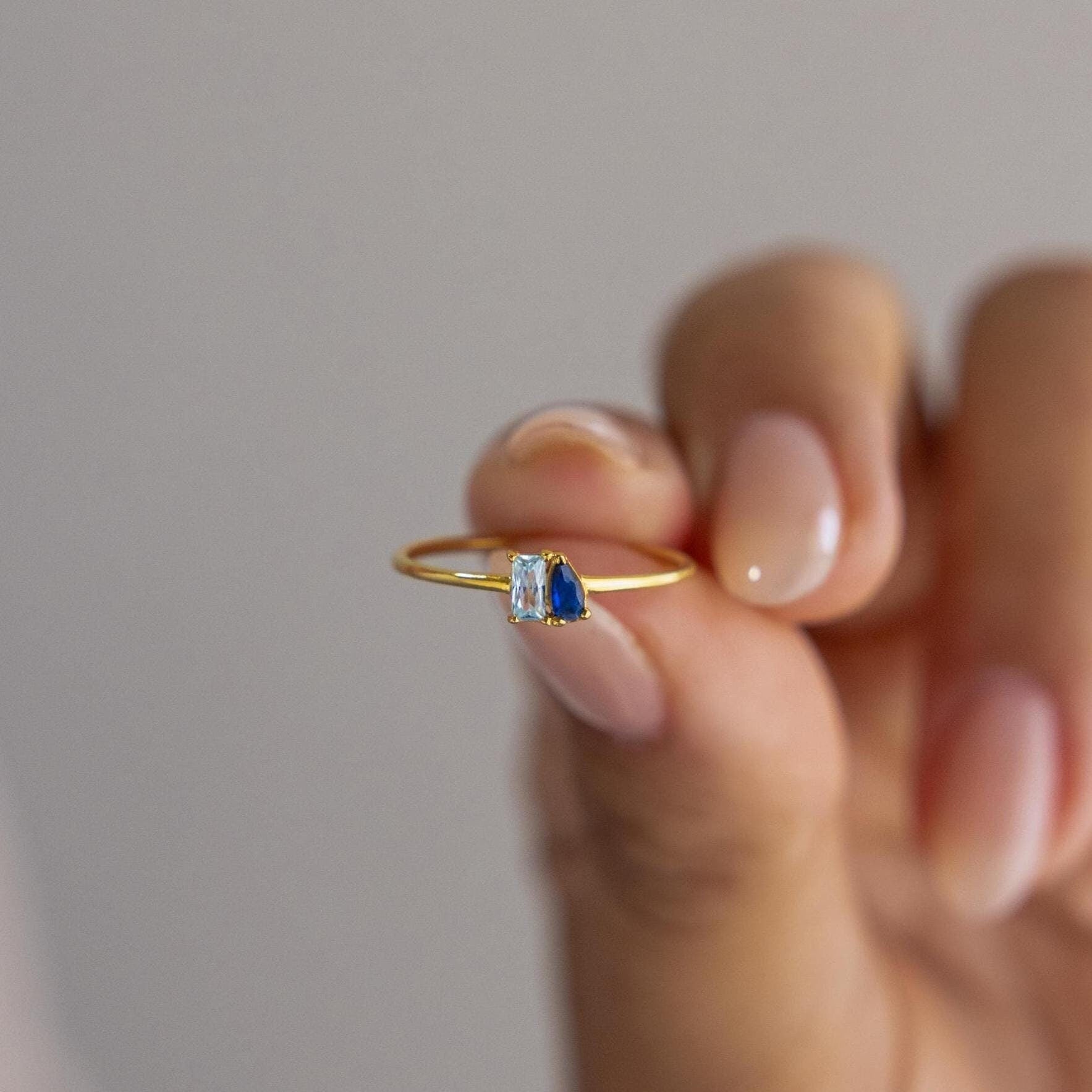A hand holds the Tiny Toi et Moi Birthstone Ring, a slim gold customizable band featuring two small gemstones—one blue and one clear.