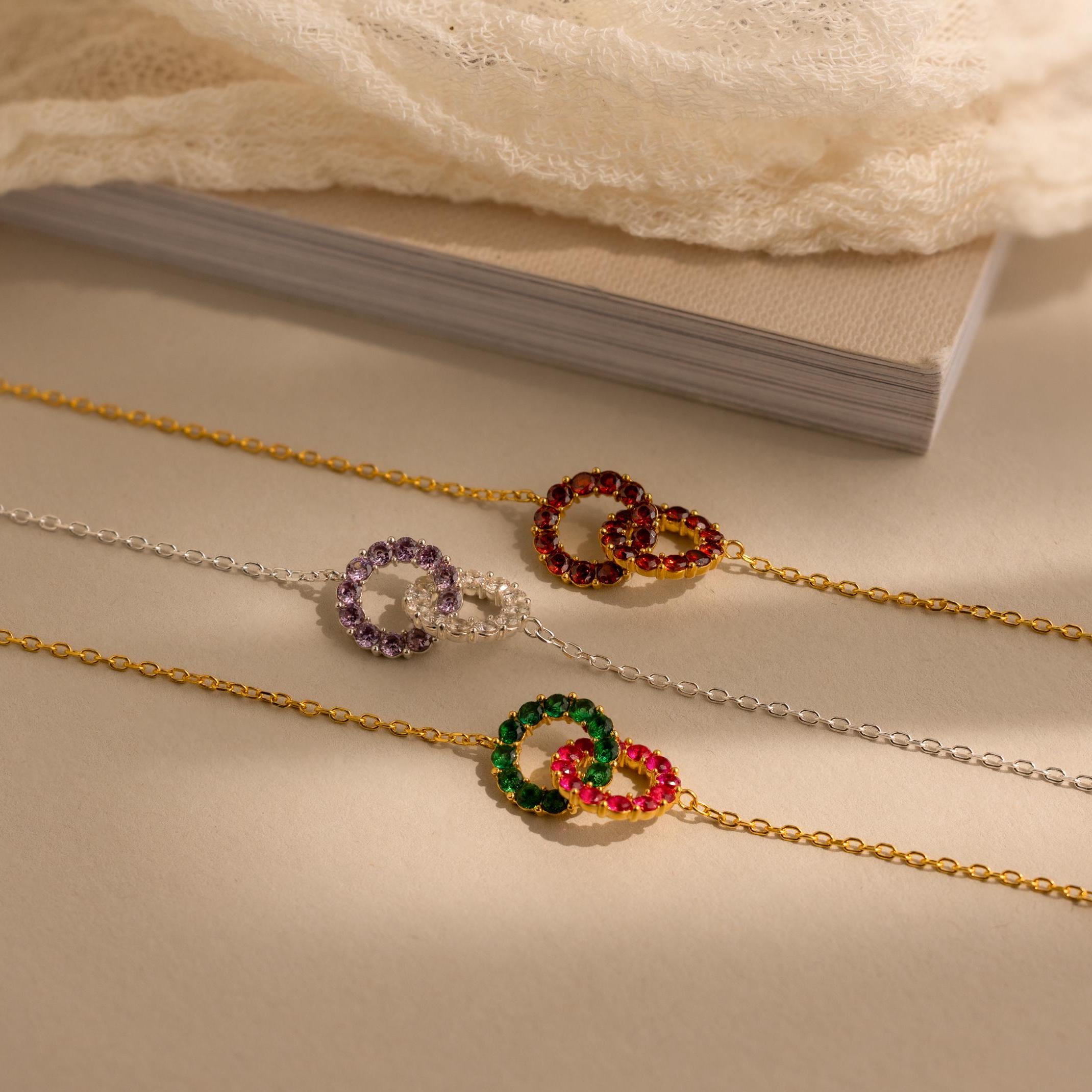 Three delicate chain bracelets with colorful interlocking circle charms rest on a beige surface by a book and lace—a thoughtful gift, perfect as the Interlocking Duo Birthstone Bracelet.