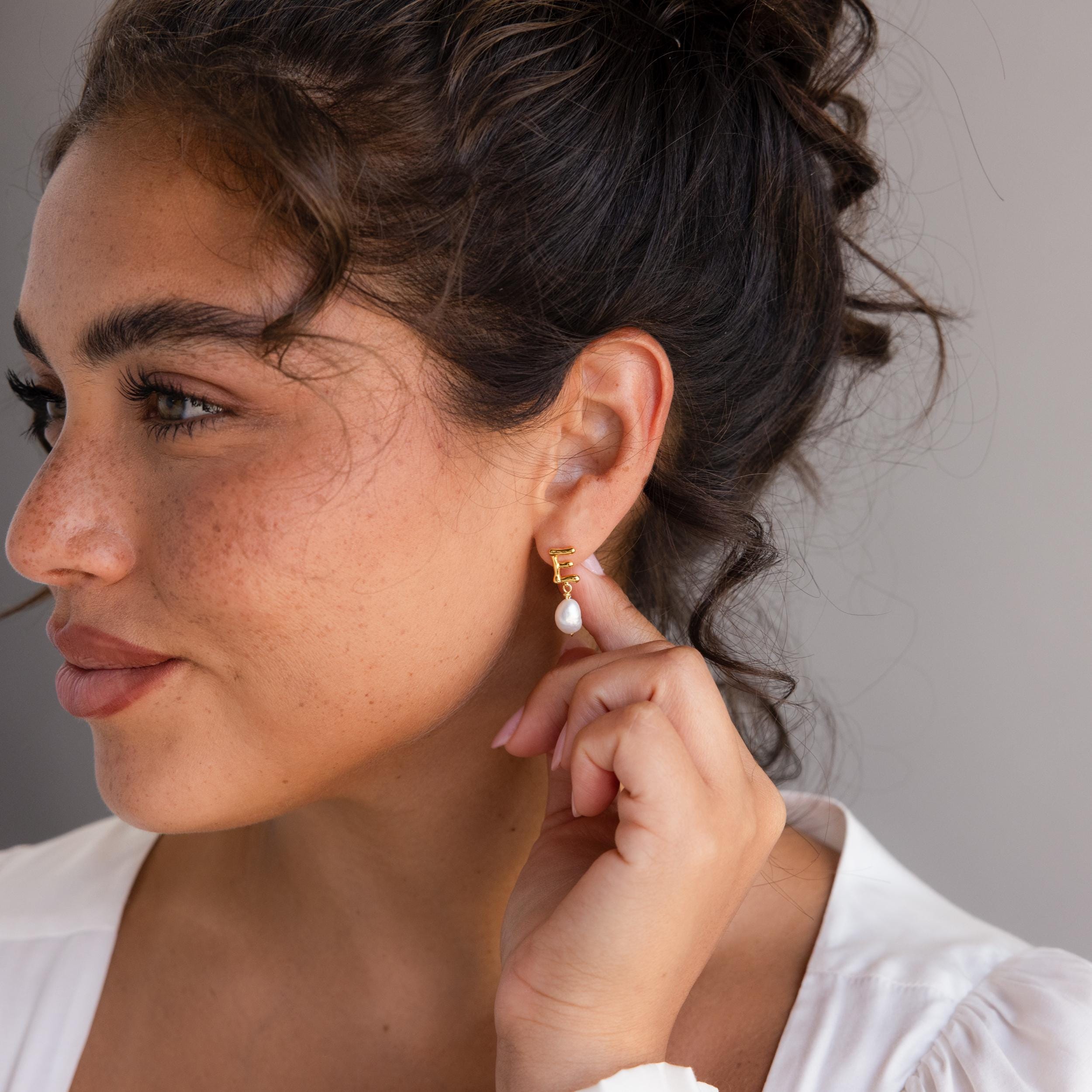 A woman with her hand on her ear, showcasing elegant Initial Pearl Drop Studs that add a touch of unique charm to her look.