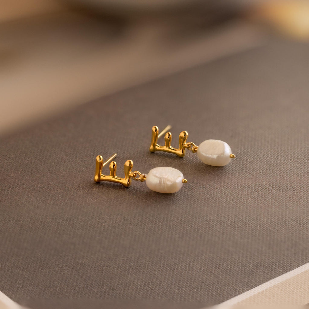 The Initial Pearl Drop Studs, featuring gold accents and white pearls with elegant Arabic calligraphy, rest on a textured brown surface.