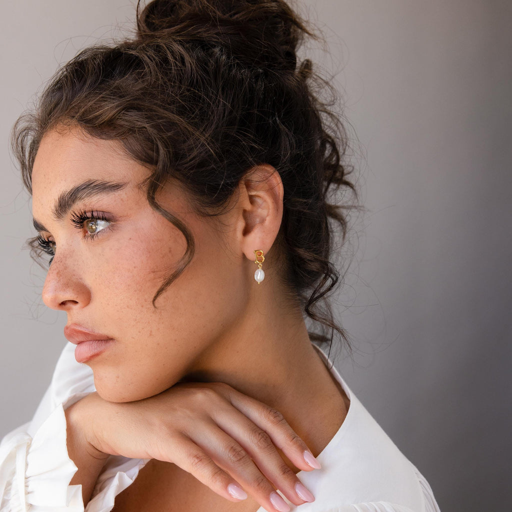 Woman with curly hair in a white top wears Initial Pearl Drop Studs, gazing thoughtfully to the side.