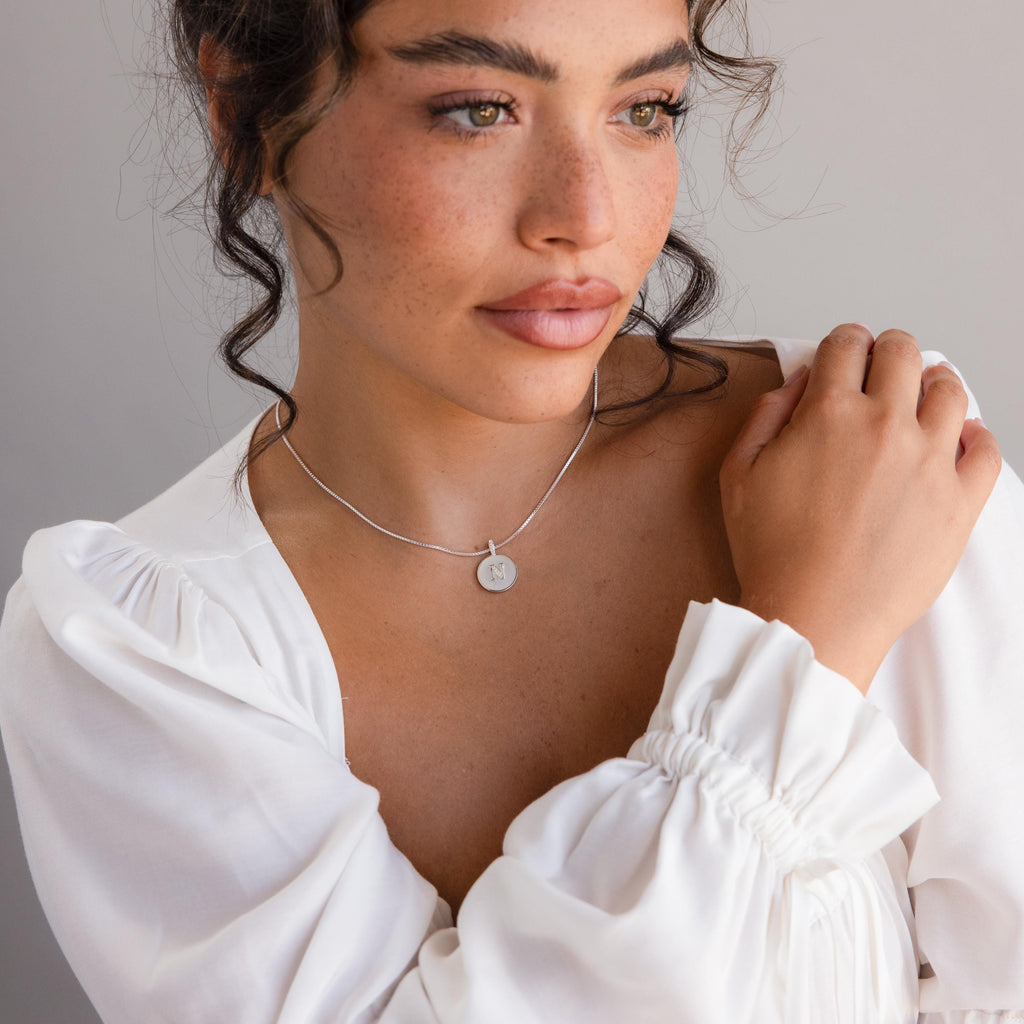 Woman with wavy hair in a white blouse, looking to the side and wearing the Gemstone Initial Disc Necklace featuring a round pendant.