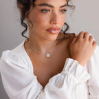 Woman with wavy hair in a white blouse, looking to the side and wearing the Gemstone Initial Disc Necklace featuring a round pendant.