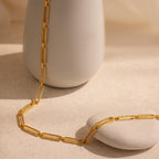 The Pave Link Chain Necklace with rectangular links is displayed draped over a smooth white vase and a stone on a beige surface.