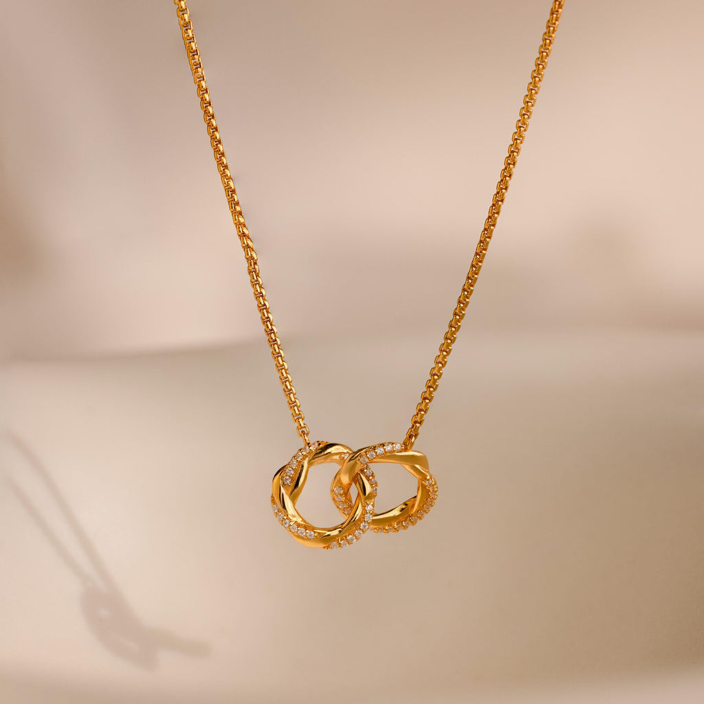 The Interlocking Pave Charm Necklace features two gold rings—one adorned with diamonds—on a soft beige background, symbolizing the timeless, unbreakable bond between mother and daughter.
