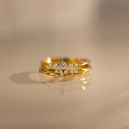 The Trio Diamond Stacking Ring Set, featuring three gold diamond rings including an art deco style, is elegantly stacked and reflected on a smooth beige surface.