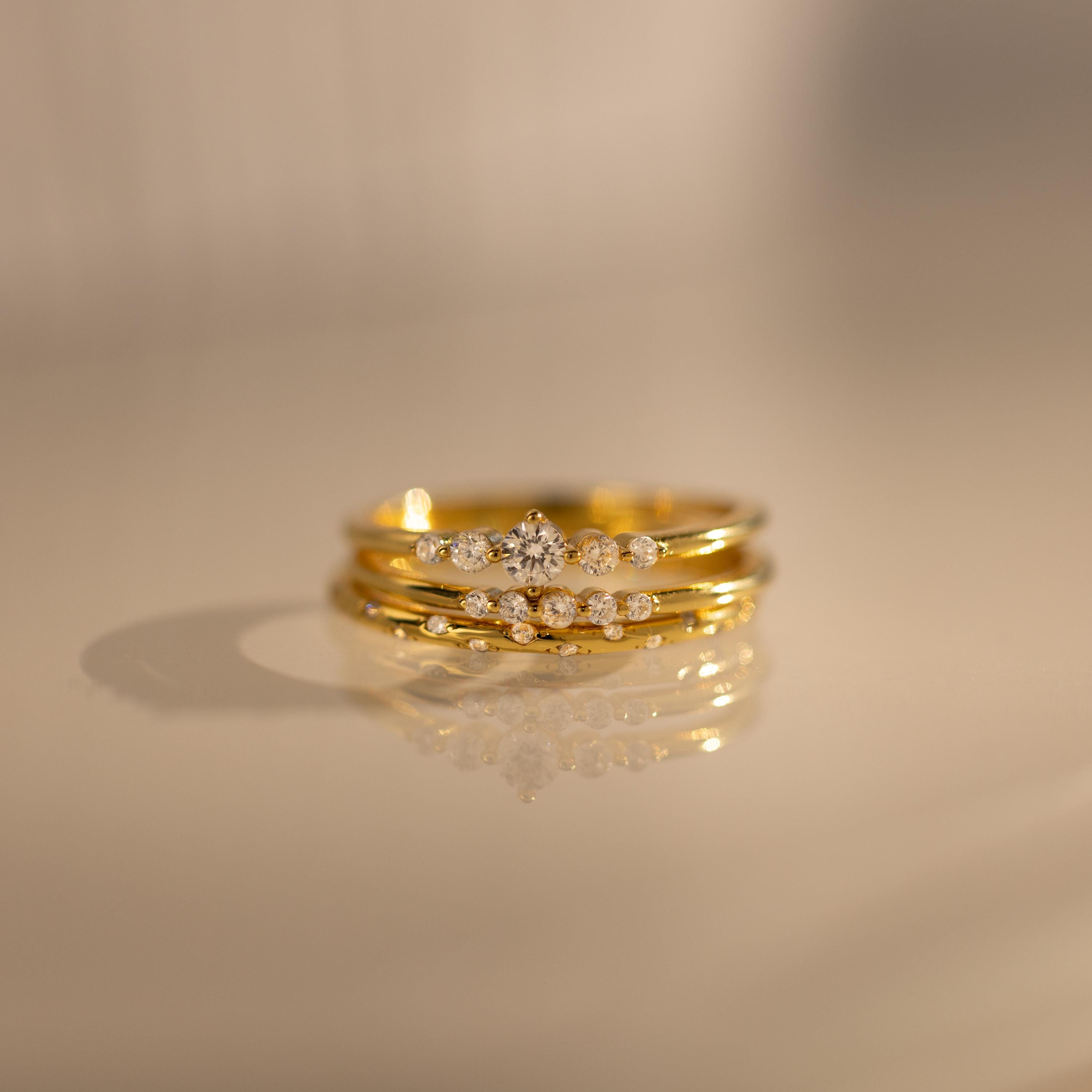 The Trio Diamond Stacking Ring Set, featuring three gold diamond rings including an art deco style, is elegantly stacked and reflected on a smooth beige surface.