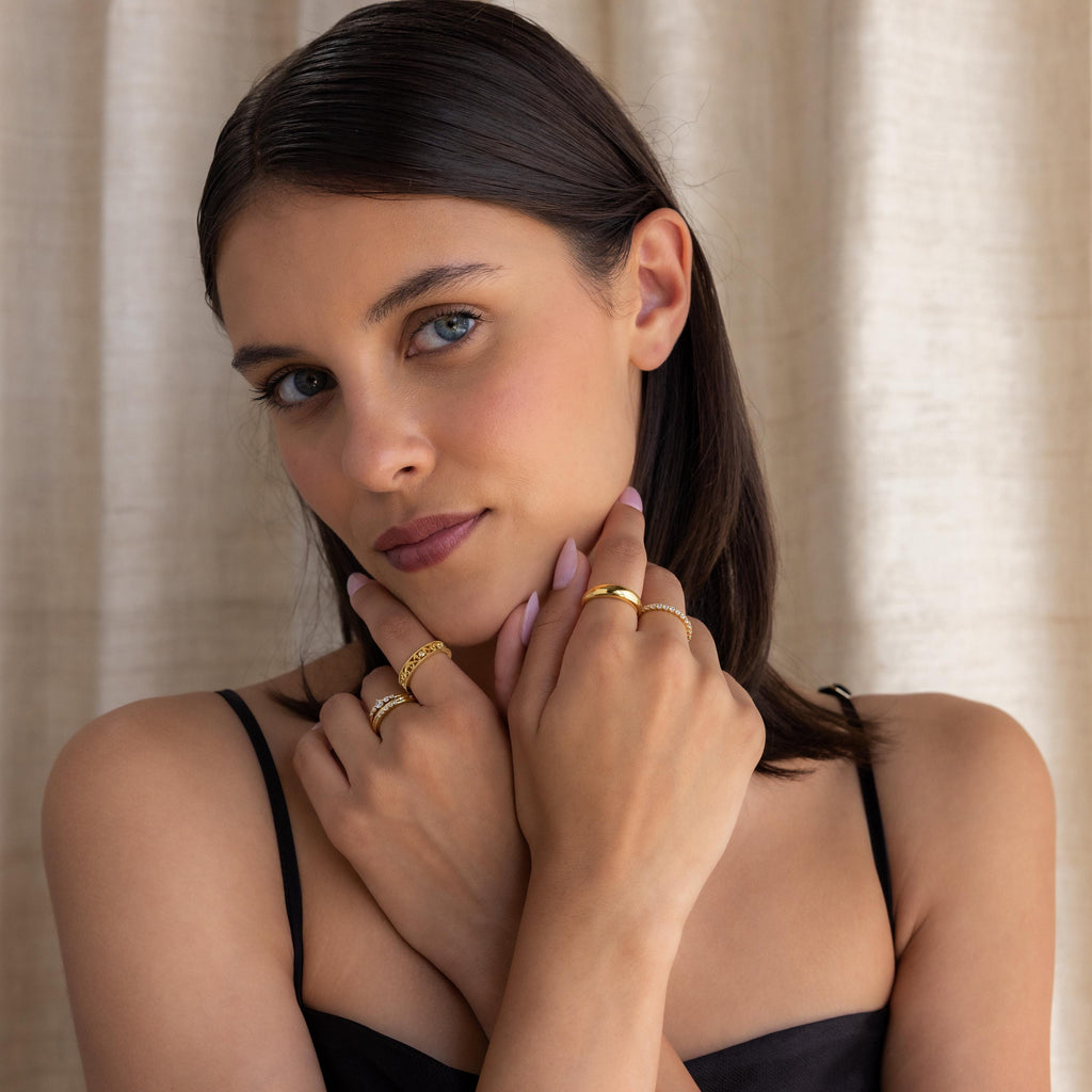 Woman with straight brown hair wears a black spaghetti strap top and gold rings, including the Trio Diamond Stacking Ring Set, as she poses with her hands near her face.