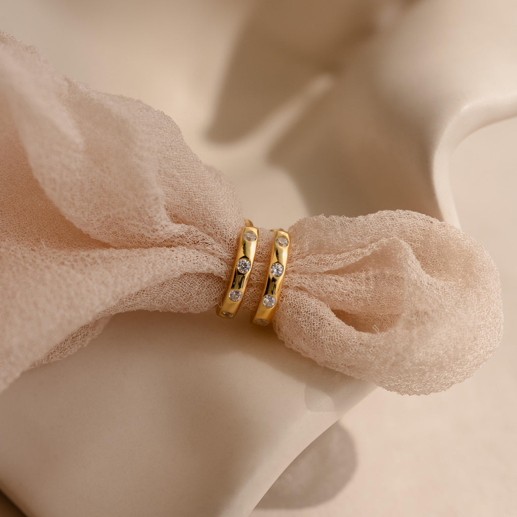 The Mini Diamond Sprinkle Huggies—two gold huggie hoops with small diamonds—are displayed on a soft blush pink textured fabric.