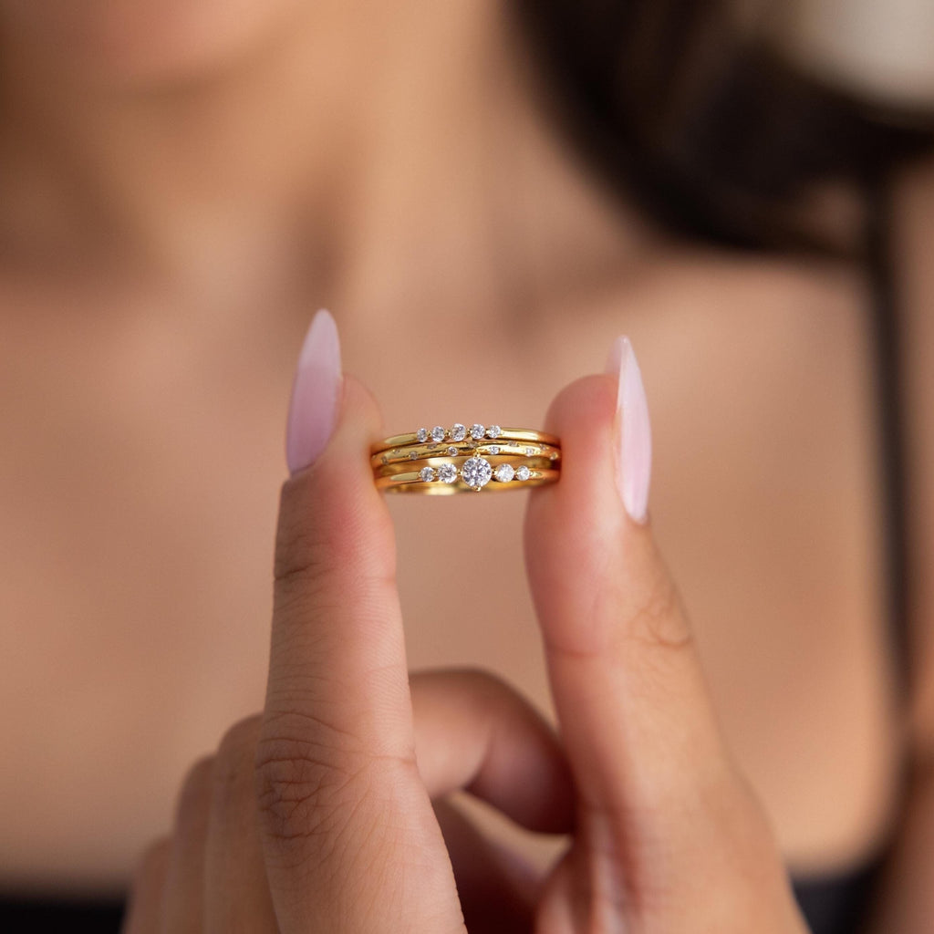 A woman holds the Trio Diamond Stacking Ring Set—featuring elegant gold and diamond rings—between her fingers, against a blurred background.