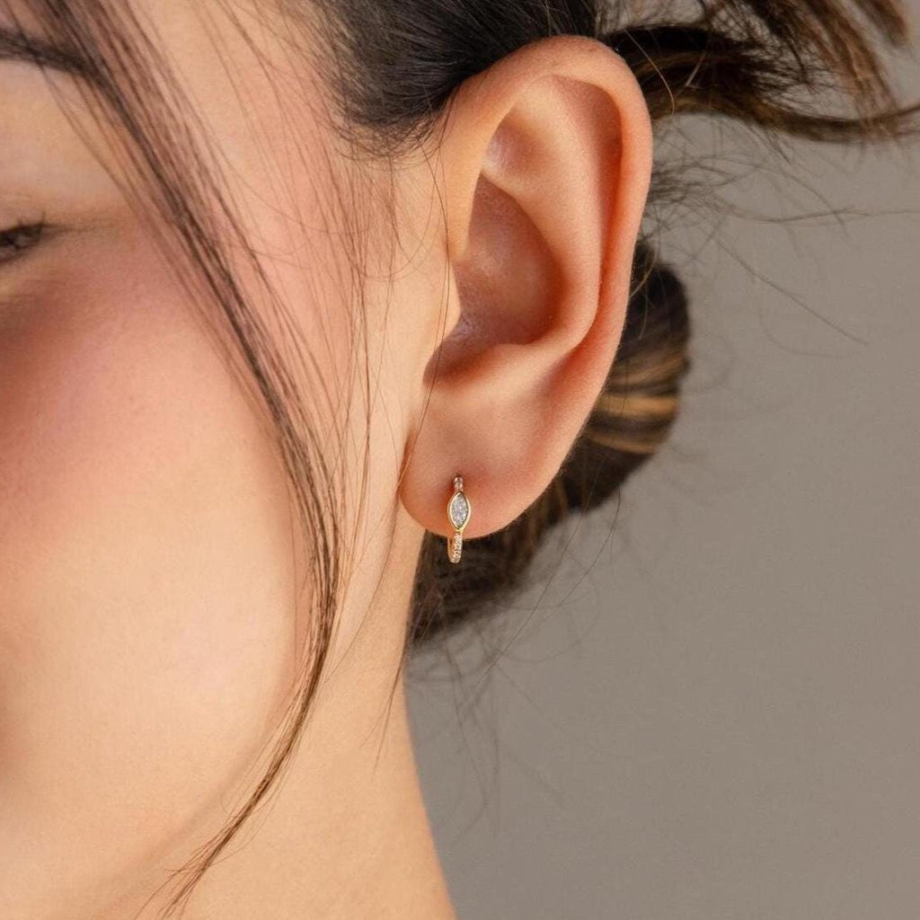 A close-up of a woman's ear wearing Marquise Pave Huggies—small gold hoop earrings featuring a bezeled marquise gemstone and art deco-inspired design.