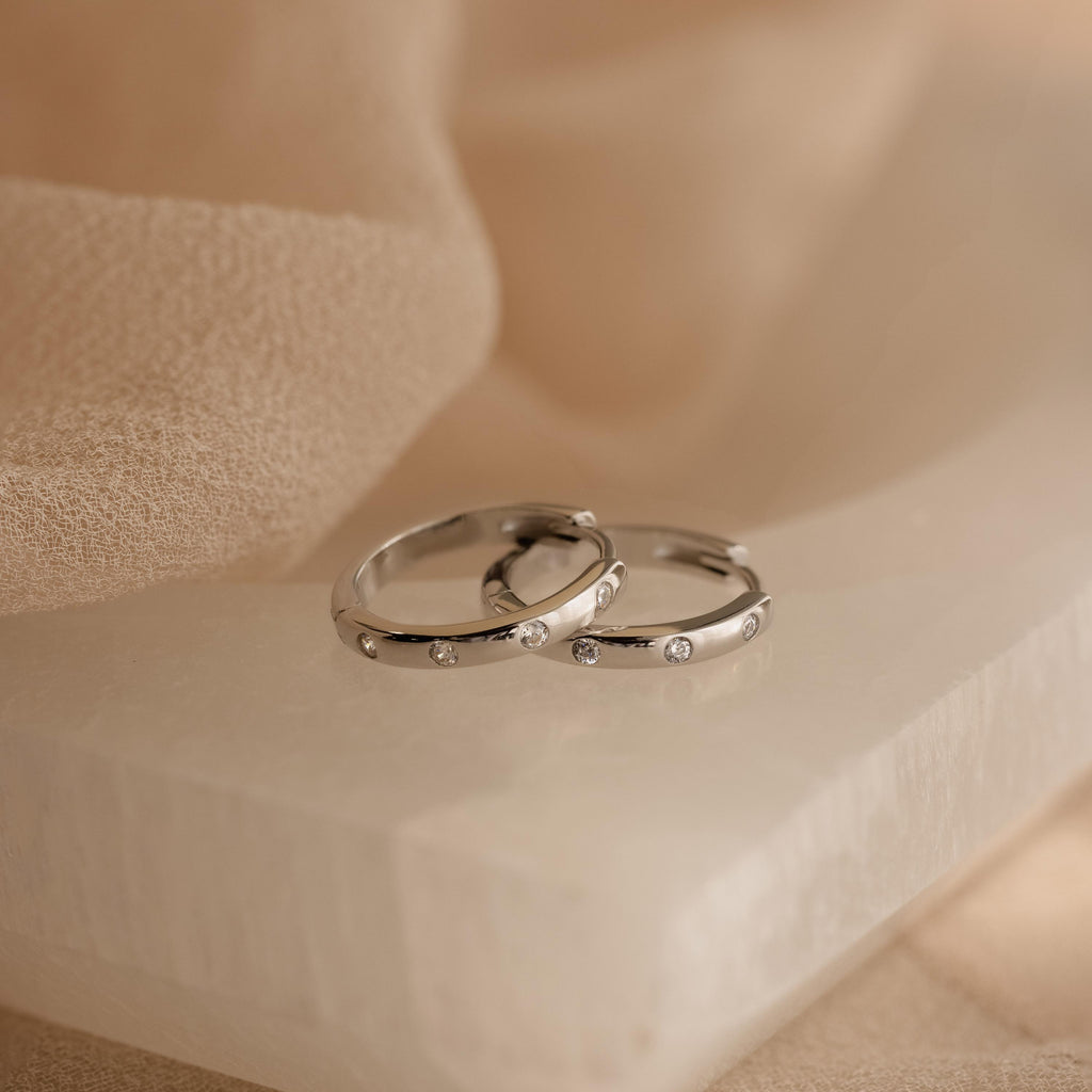 Two silver rings adorned with small diamonds rest on a smooth white stone, set against soft beige fabric—an elegant tribute to the Classic Diamond Sprinkle Huggies.