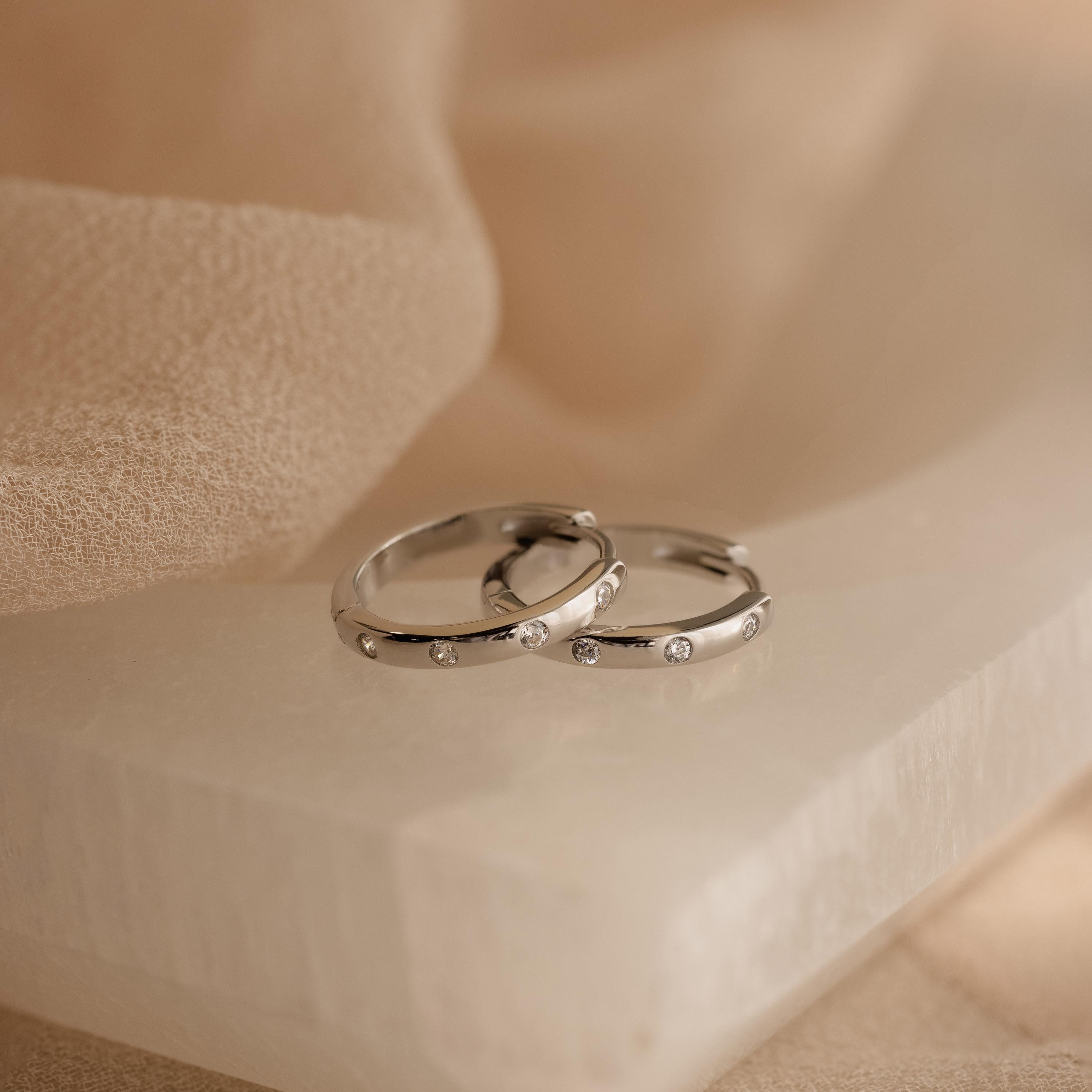Two silver rings adorned with small diamonds rest on a smooth white stone, set against soft beige fabric—an elegant tribute to the Classic Diamond Sprinkle Huggies.